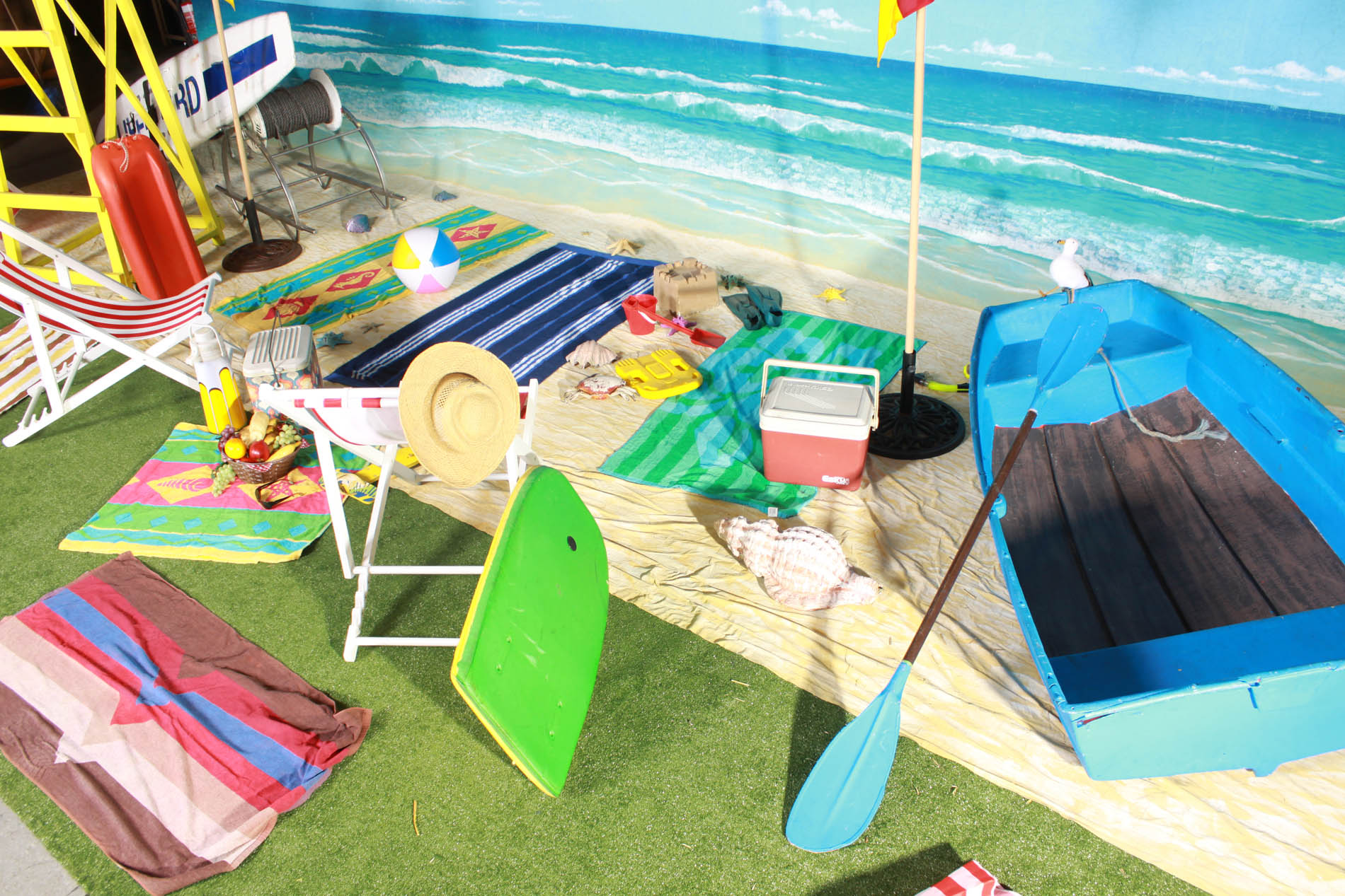 Beach day set with striped towels, deck chairs, a green boogie board, esky, beach ball and a blue timber rowboat against a painted ocean backdrop