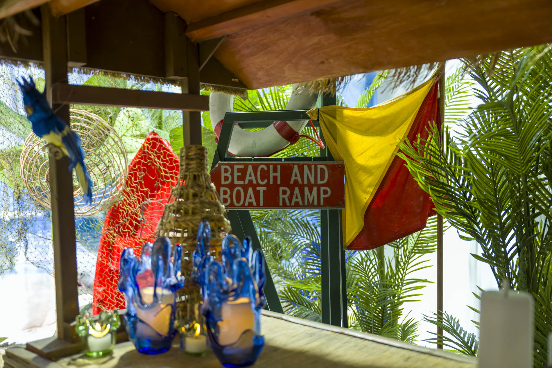 Thatched beach shack bar with a Beach and Boat Ramp sign, signal flag, life ring and blue glass candle vessels
